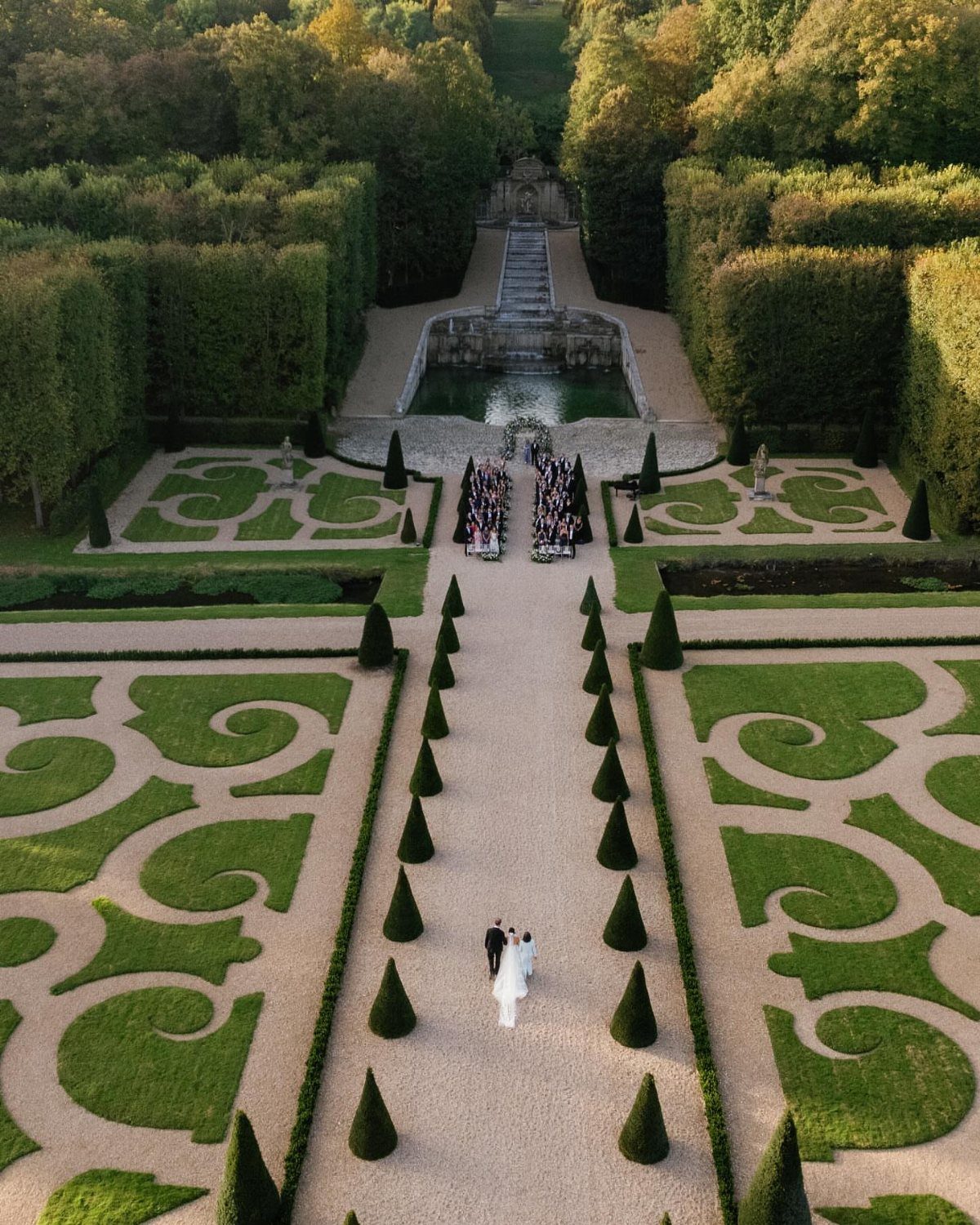 Bride and groom walking through a formal garden with symmetrical hedges and fountains