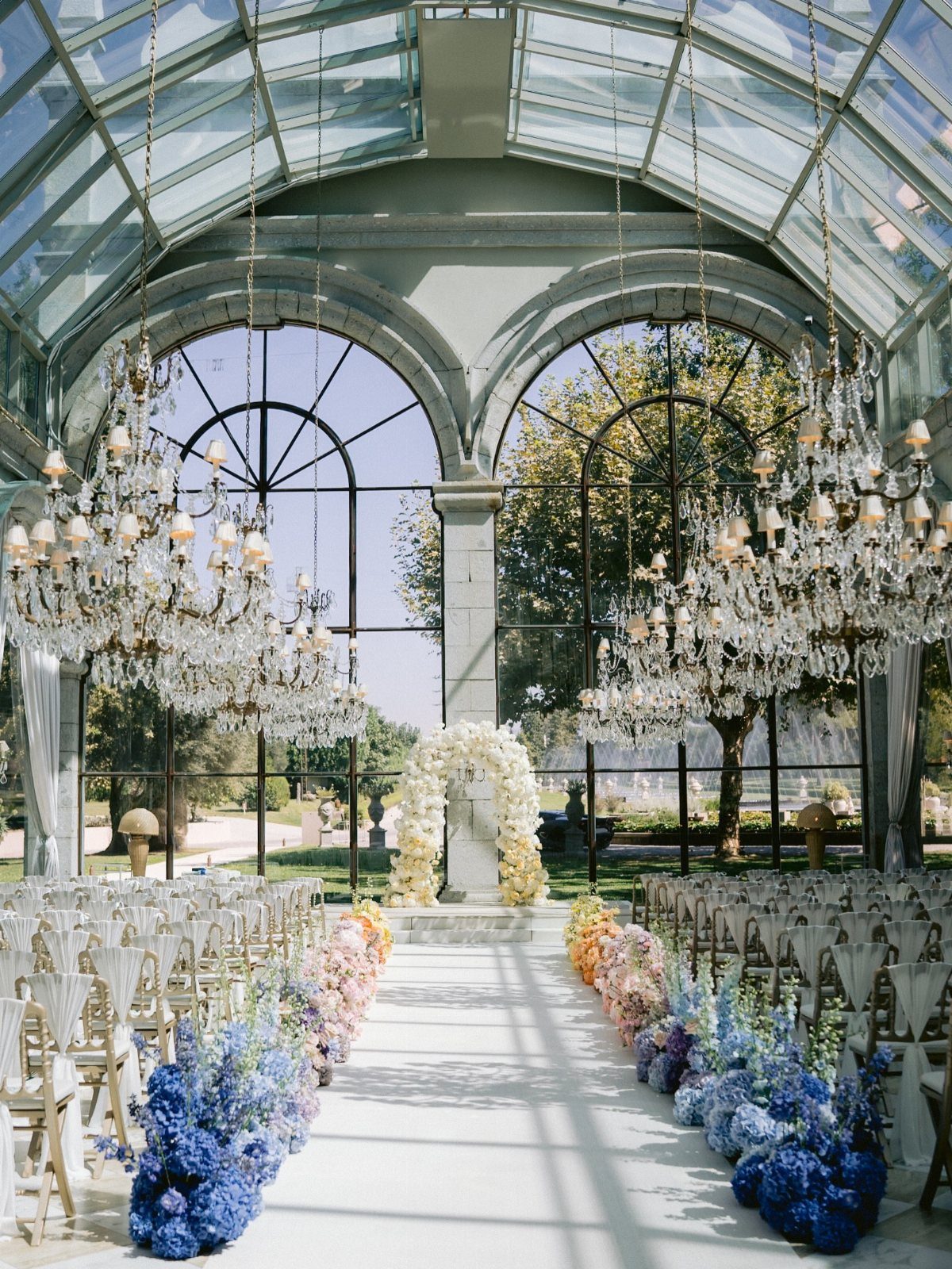Wedding ceremony setup in a glass conservatory with crystal chandeliers and floral aisle.