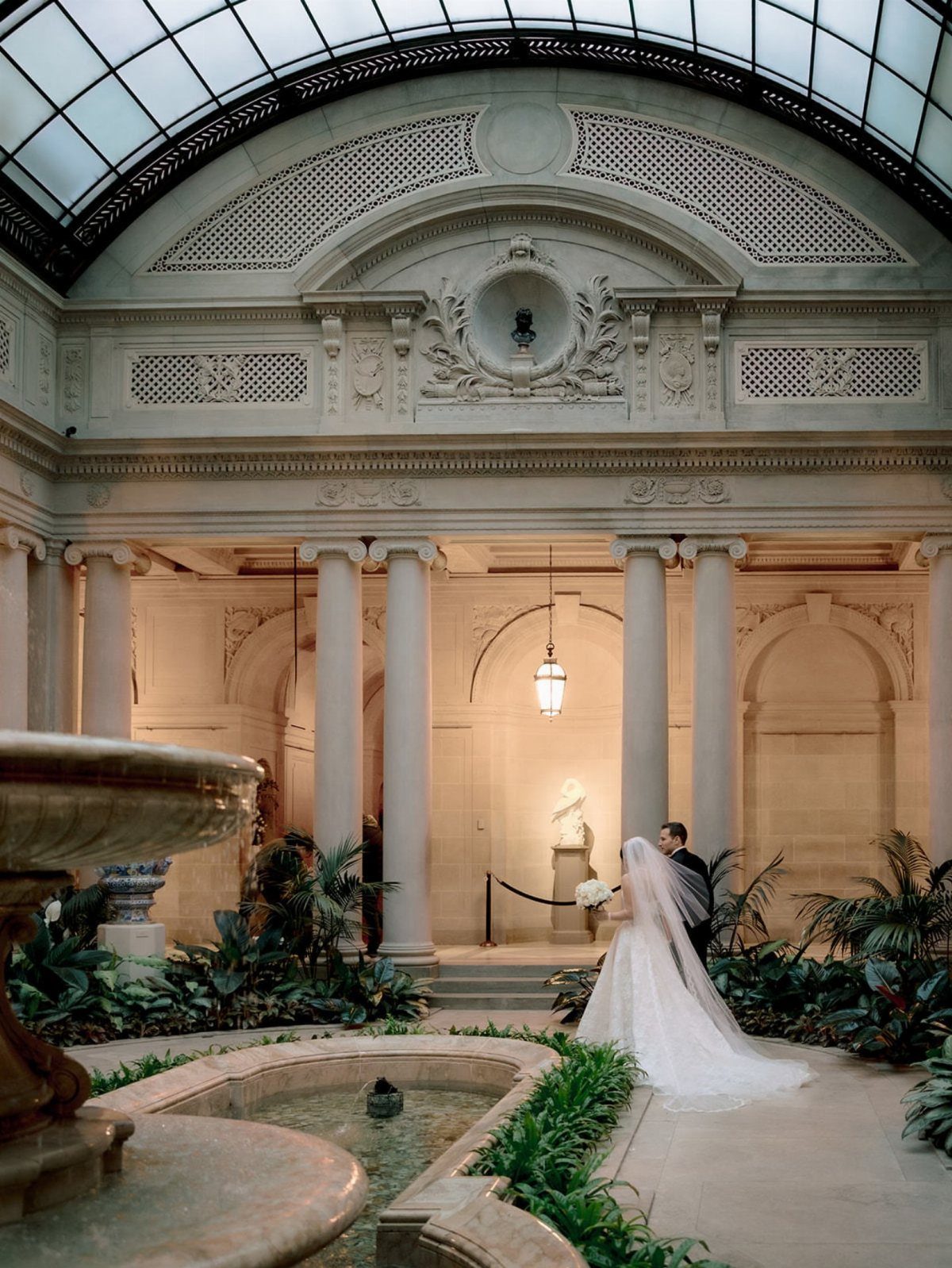 Bride and groom standing in an ornate indoor courtyard with columns and glass ceiling.