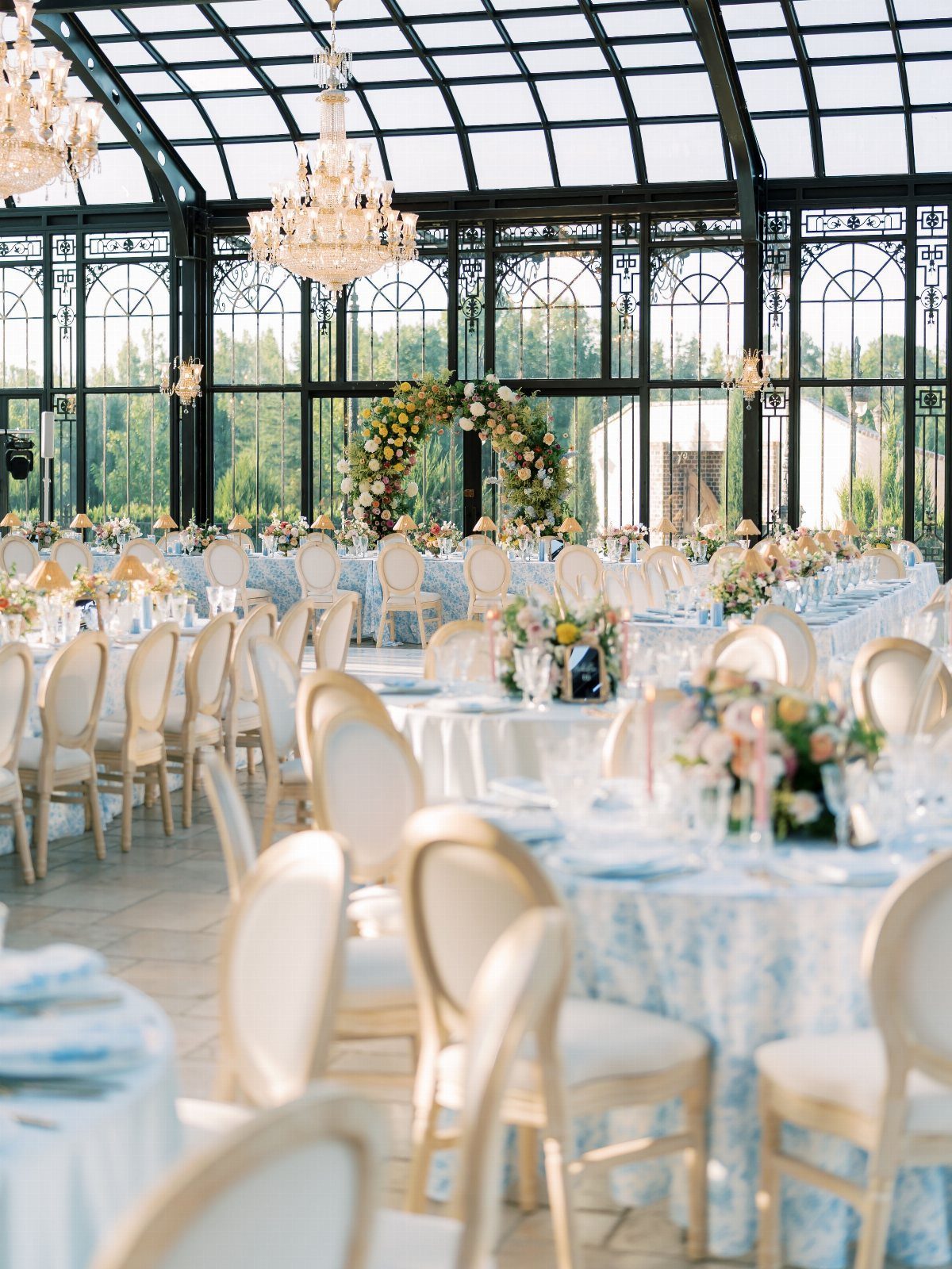 Elegant wedding reception setup in a glass conservatory with chandeliers and floral arrangements
