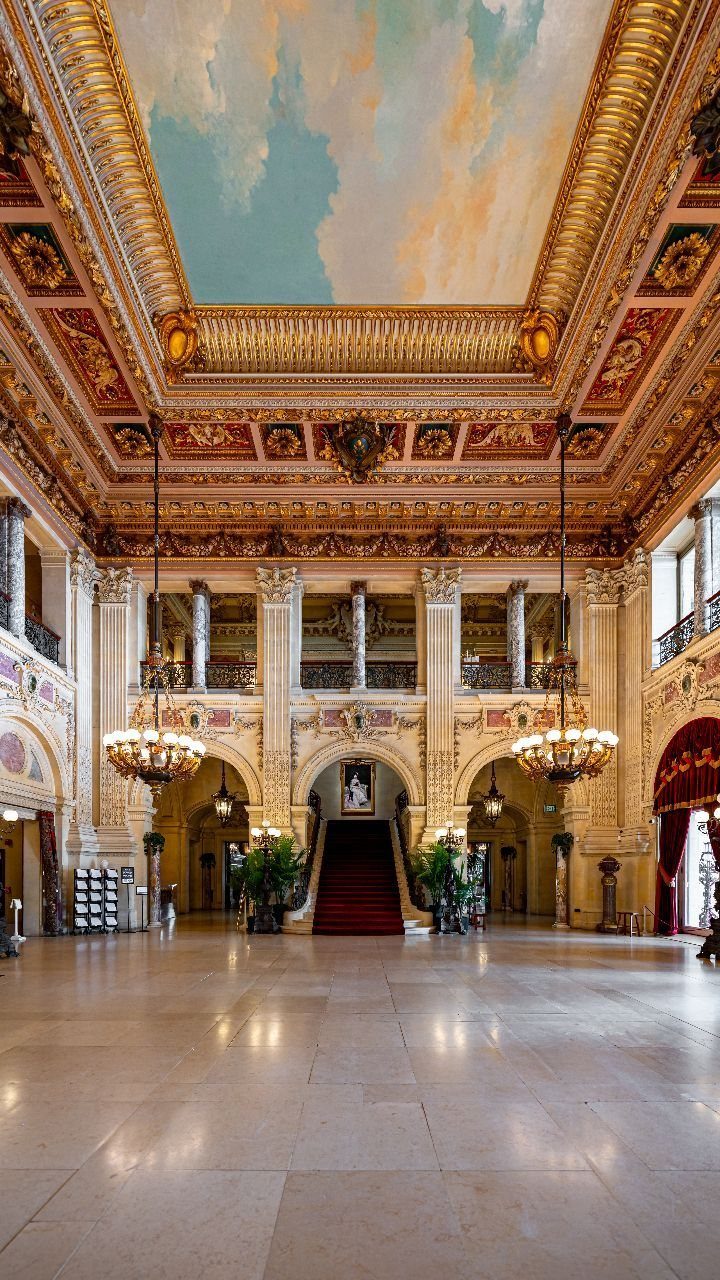 Grand hotel lobby with gold ornate ceilings, chandeliers, and a red carpet staircase.