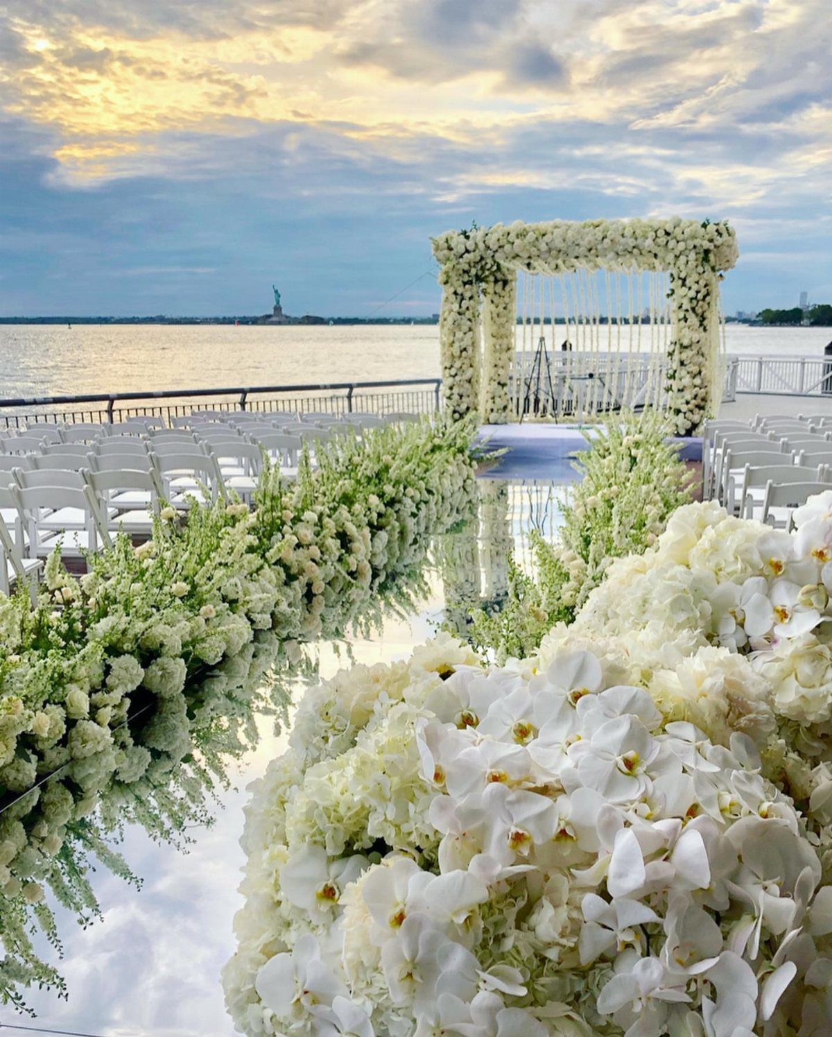 Outdoor wedding ceremony setup on a pier with white flowers and Statue of Liberty background
