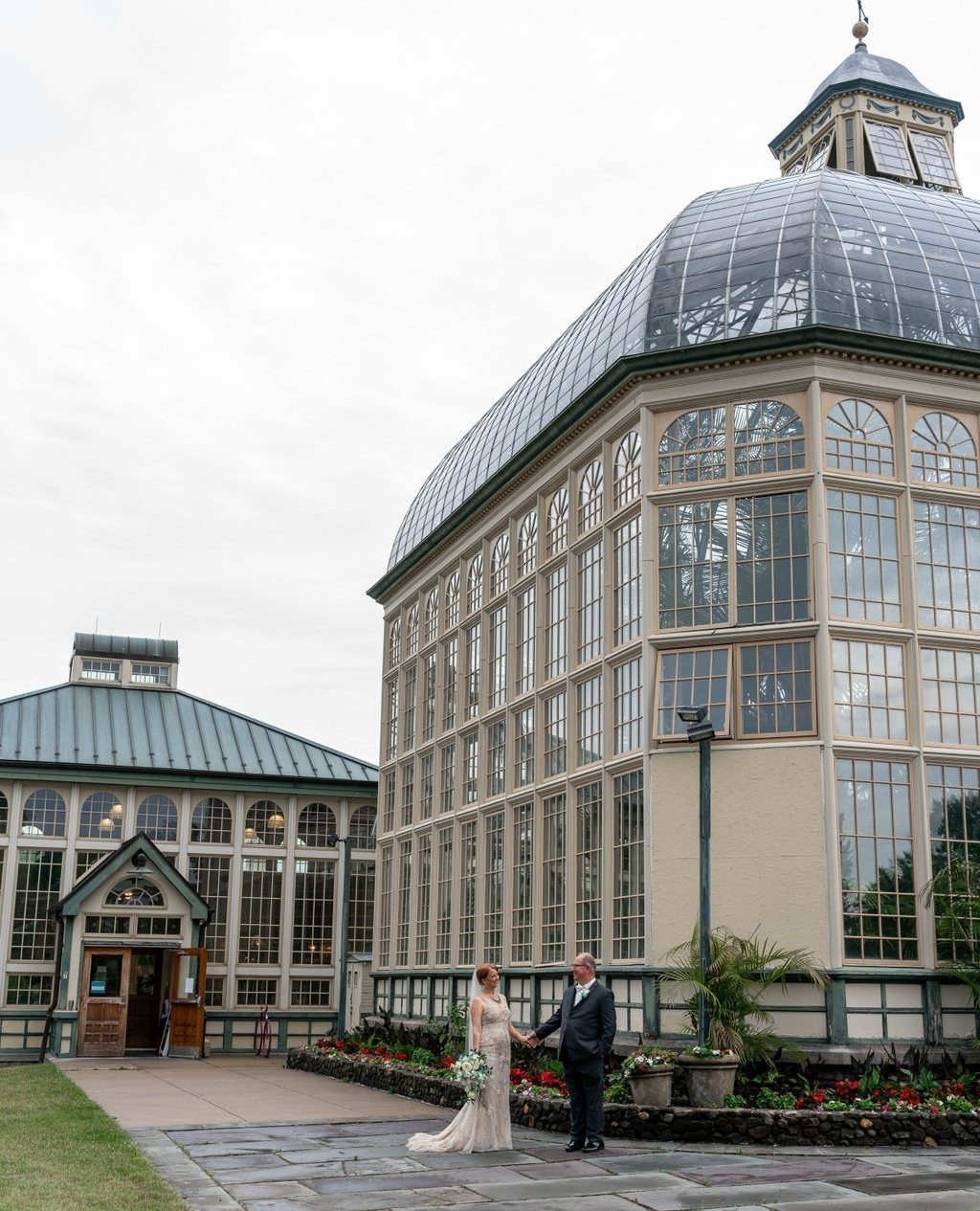 Bride and groom holding hands before a large glass conservatory wedding venue.