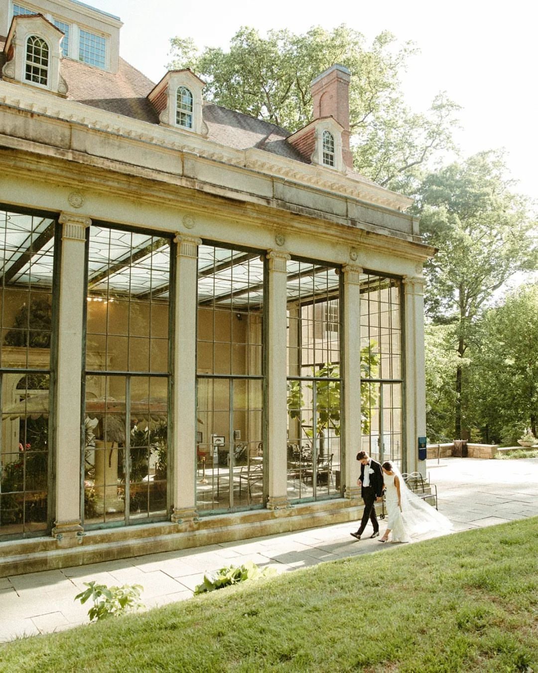 Bride and groom walking past a historic stone building with large glass windows.