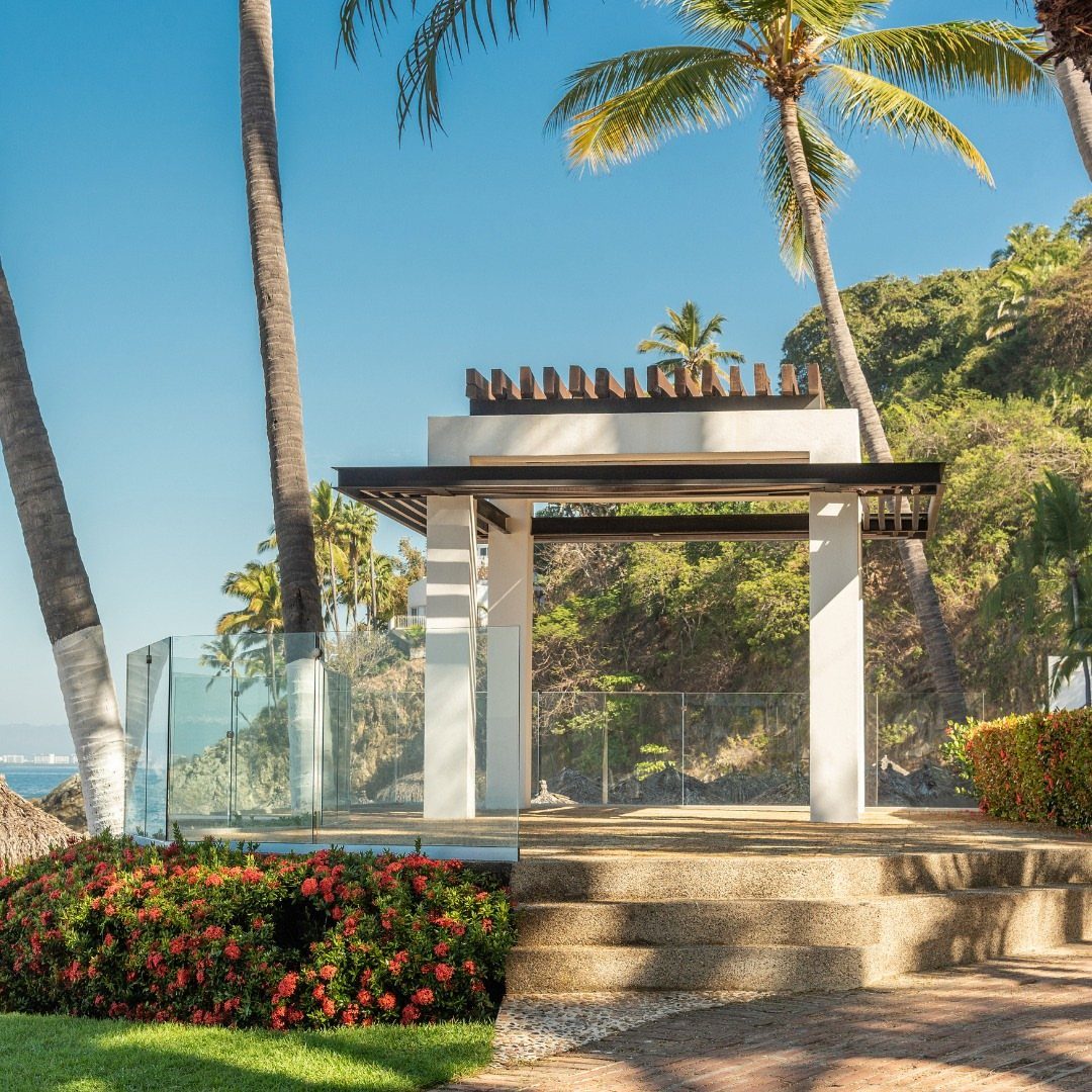 Modern white outdoor pavilion with wooden pergola roof surrounded by palm trees and ocean.