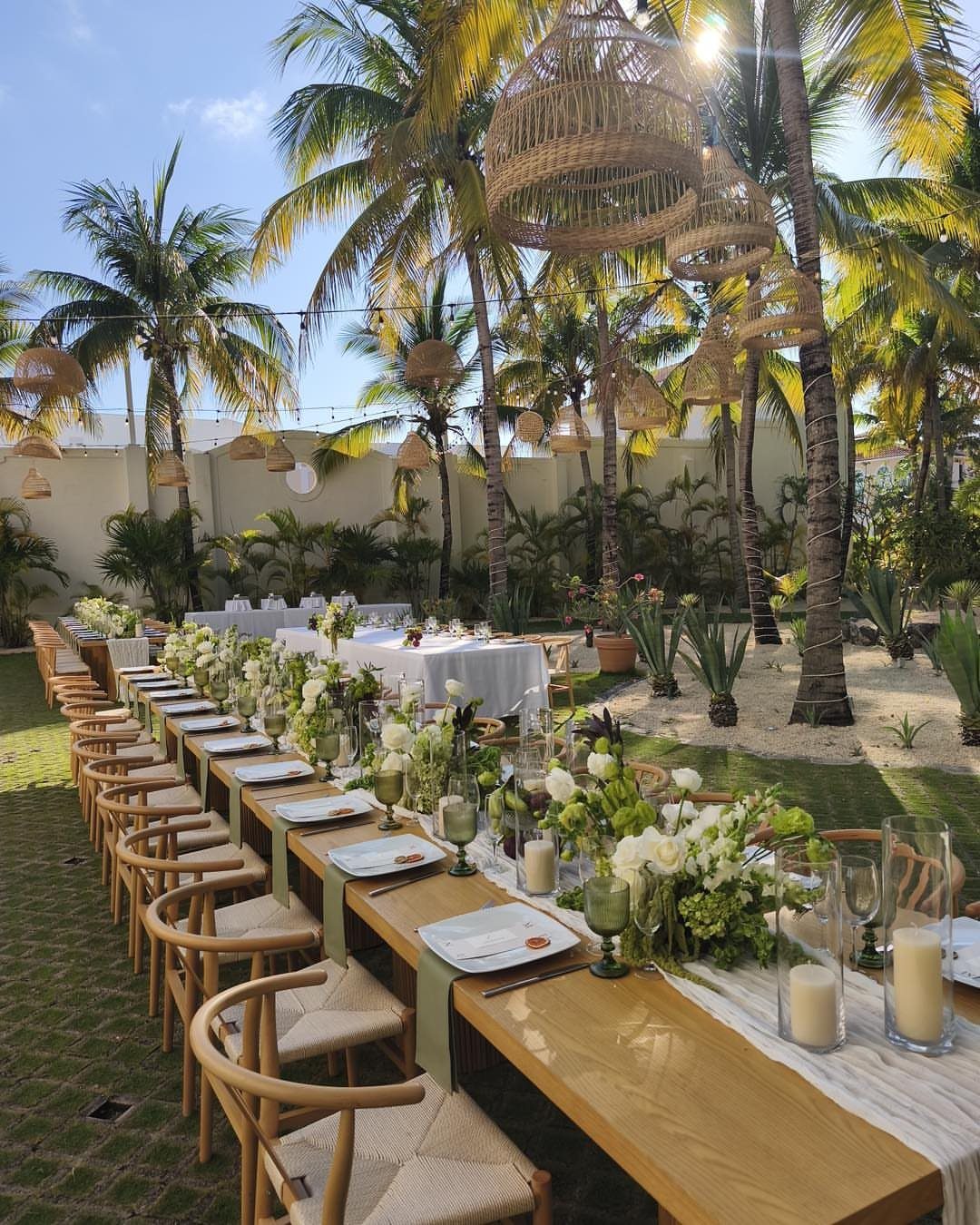 Long wooden wedding table with white flowers and wicker lamps under palm trees.