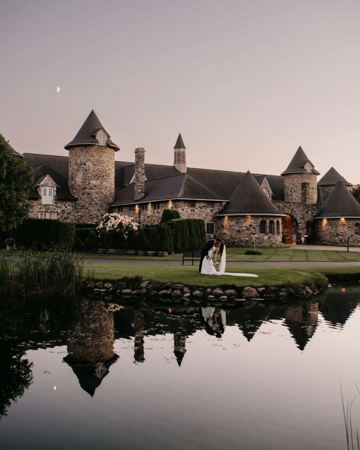 Bride and groom embracing before a stone castle estate reflected in a pond.