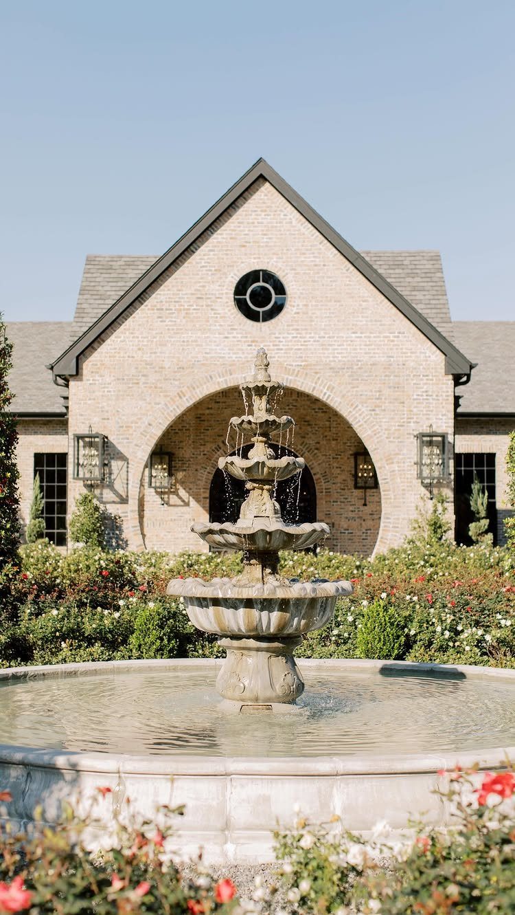 Tiered stone fountain in front of a brick building with rose gardens.