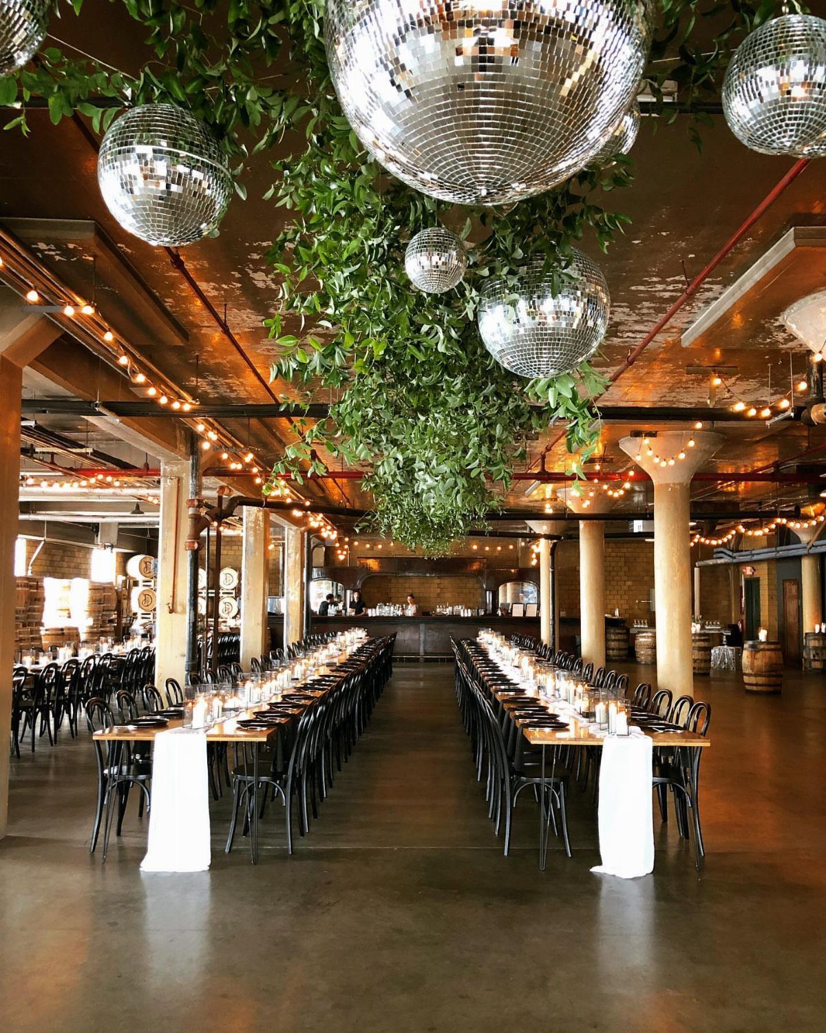 Industrial event space with long wooden tables, hanging disco balls, and greenery.