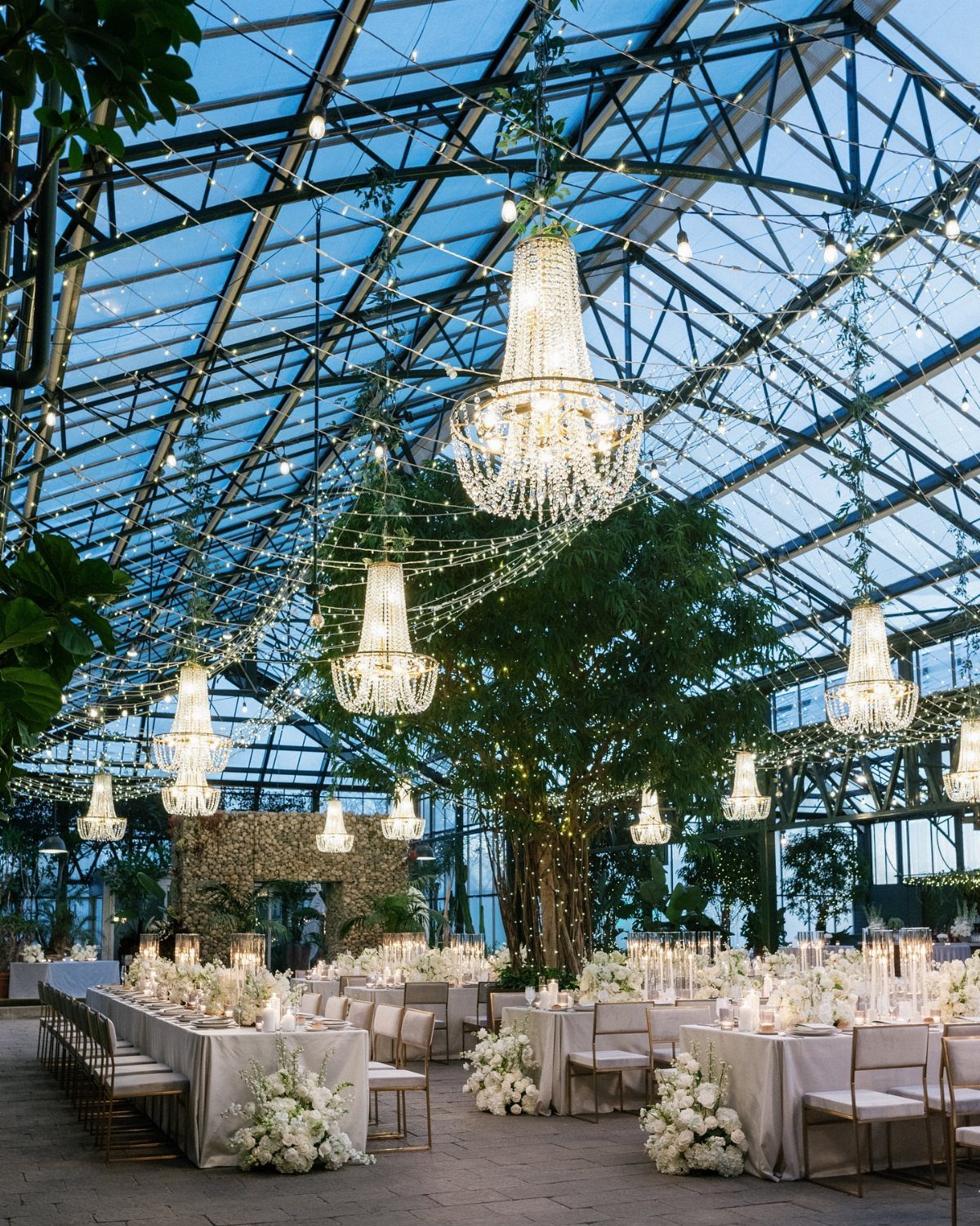 Glass conservatory wedding venue with crystal chandeliers, white tables, and lush greenery.