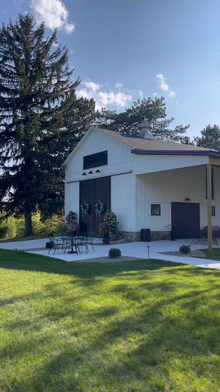 White barn wedding venue with a paved patio and tall evergreen trees.