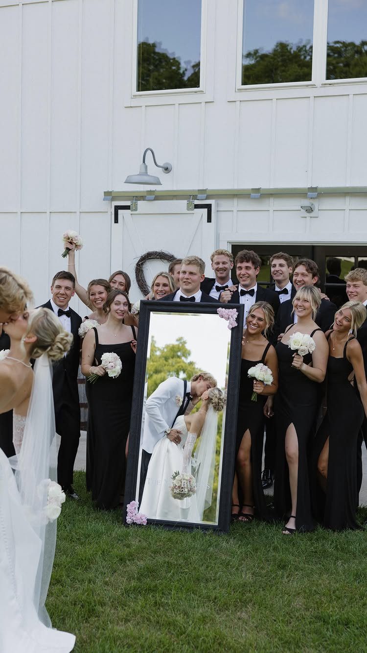 Wedding party posing outdoors with a mirror reflecting the kissing bride and groom.