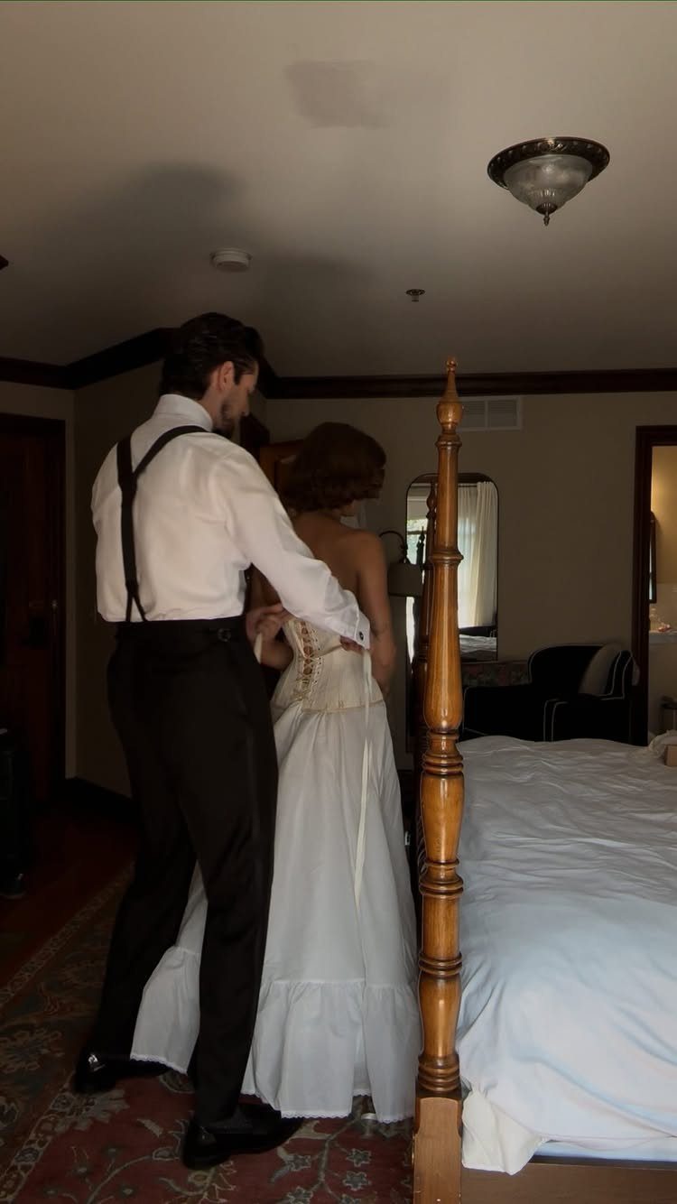 Groom in suspenders helping bride fasten the back of her white wedding dress.