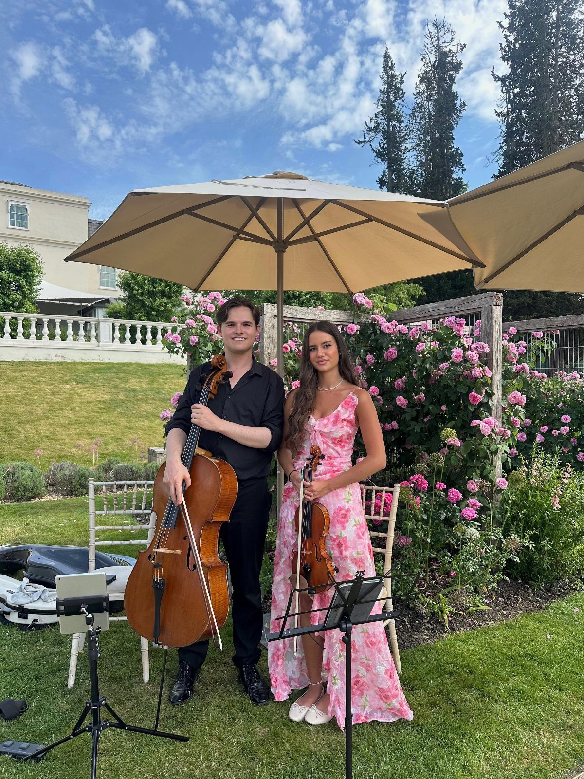 Musicians with a cello and violin posing under umbrellas in a garden setting.