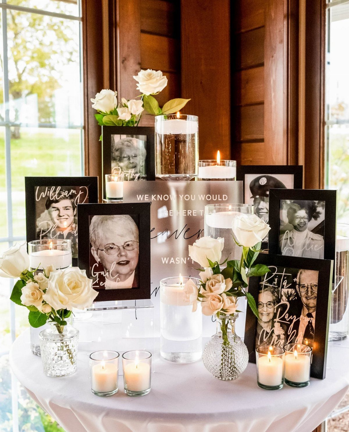 Memory table with framed portraits, white roses, and lit candles on a white cloth.