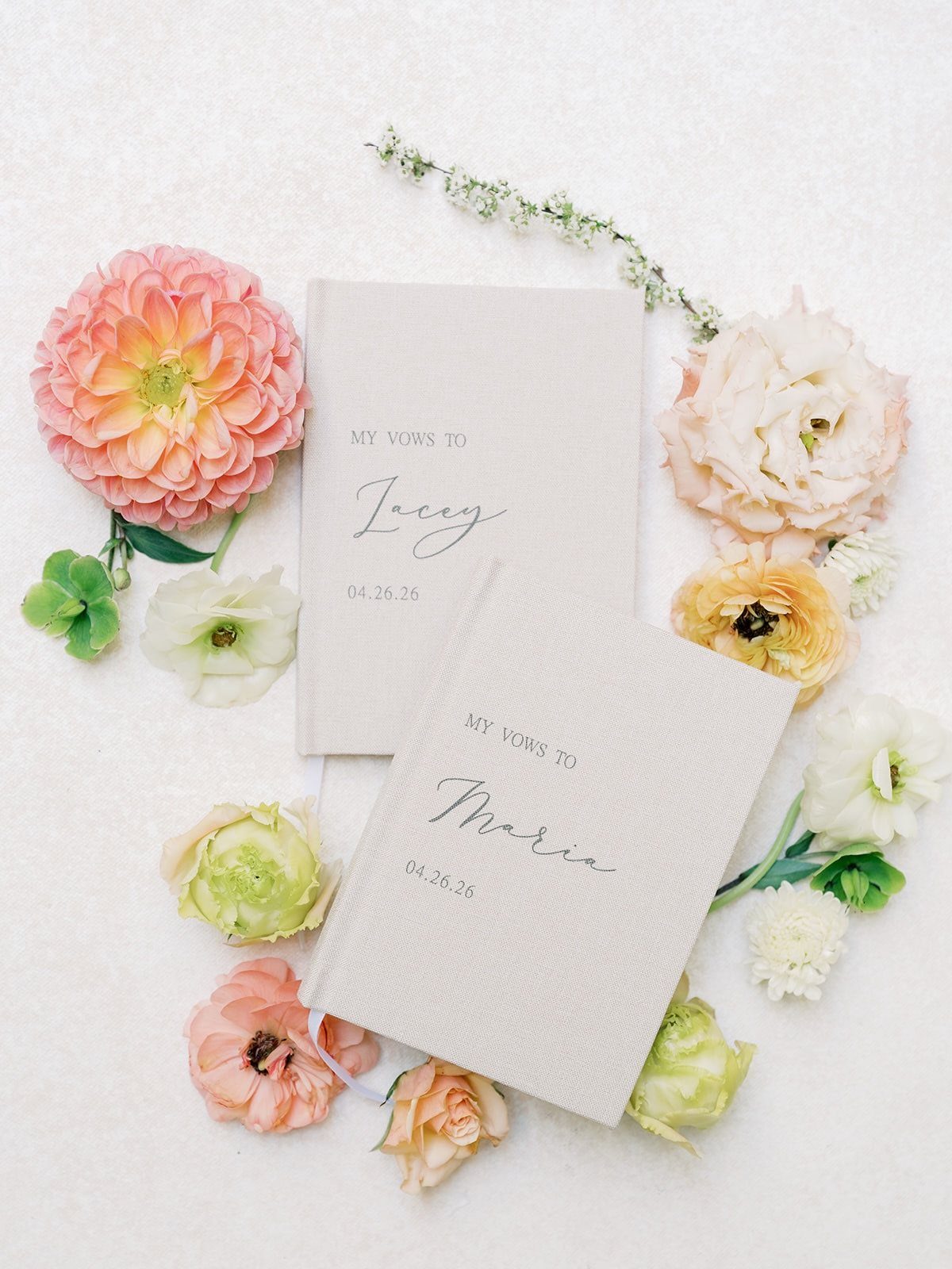 Two beige linen vow books surrounded by colorful spring flowers on white background