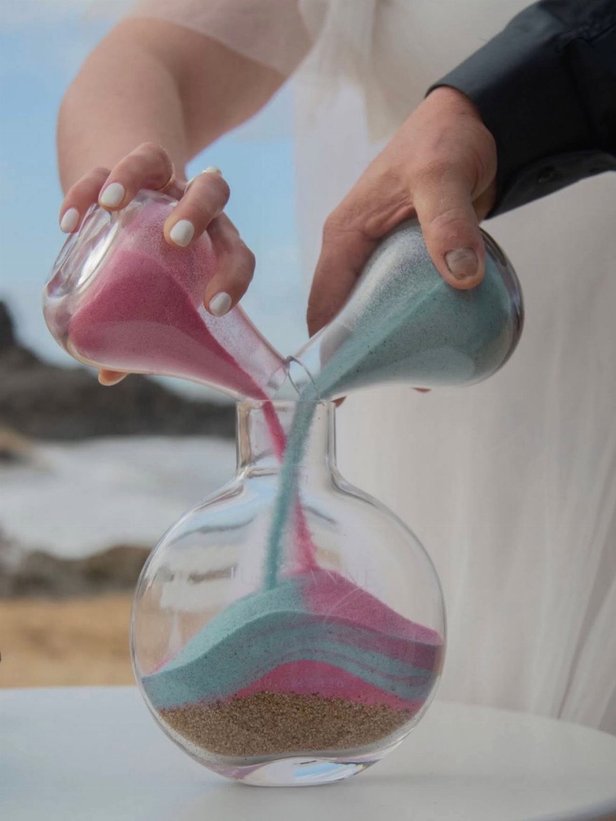 Hands pouring pink and blue sand into a shared glass vessel on a beach.