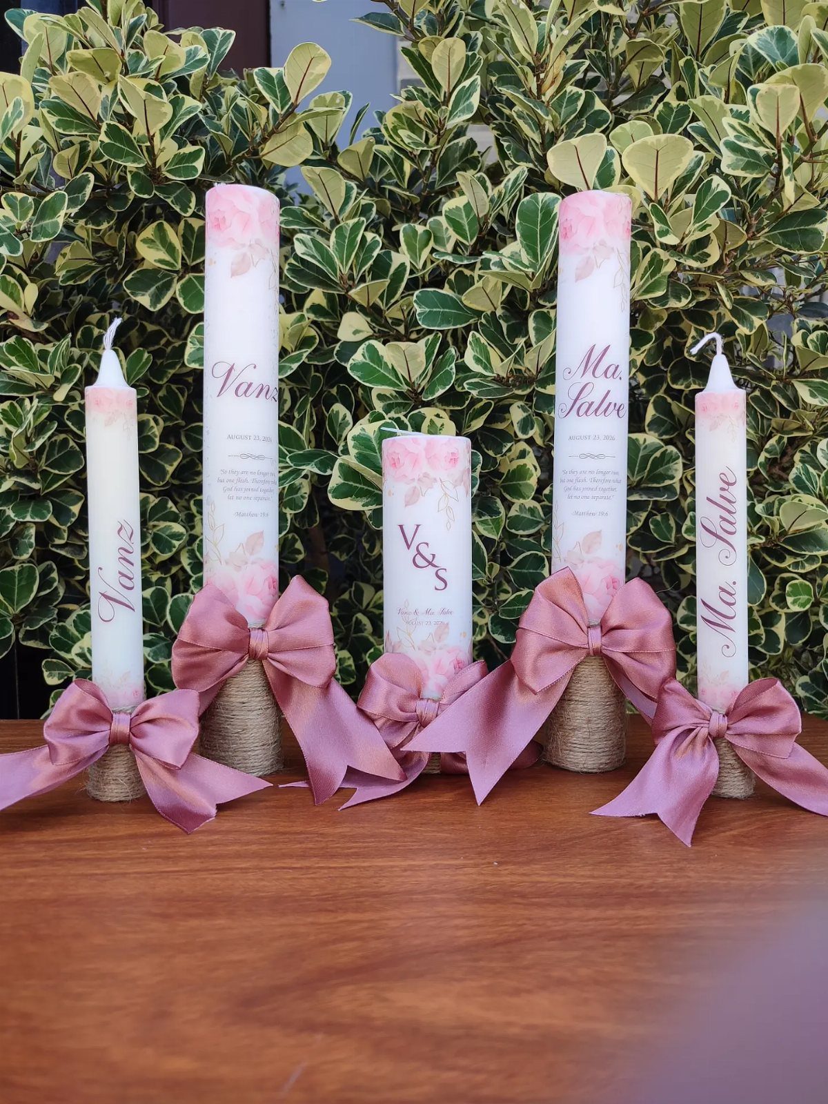 Five white wedding unity candles with pink bows and floral designs on wood.