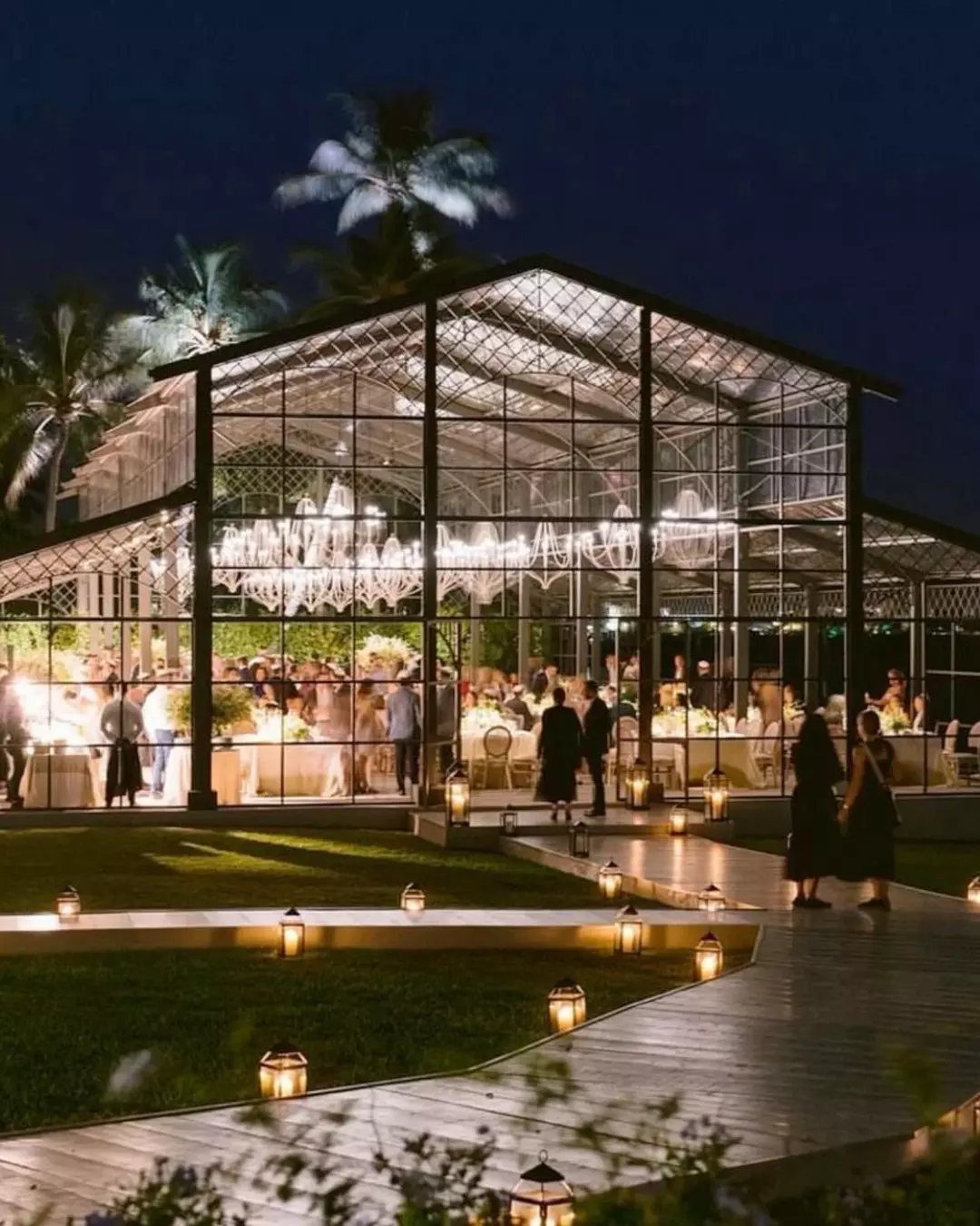 Illuminated glass house event venue at night with lanterns on a wooden walkway