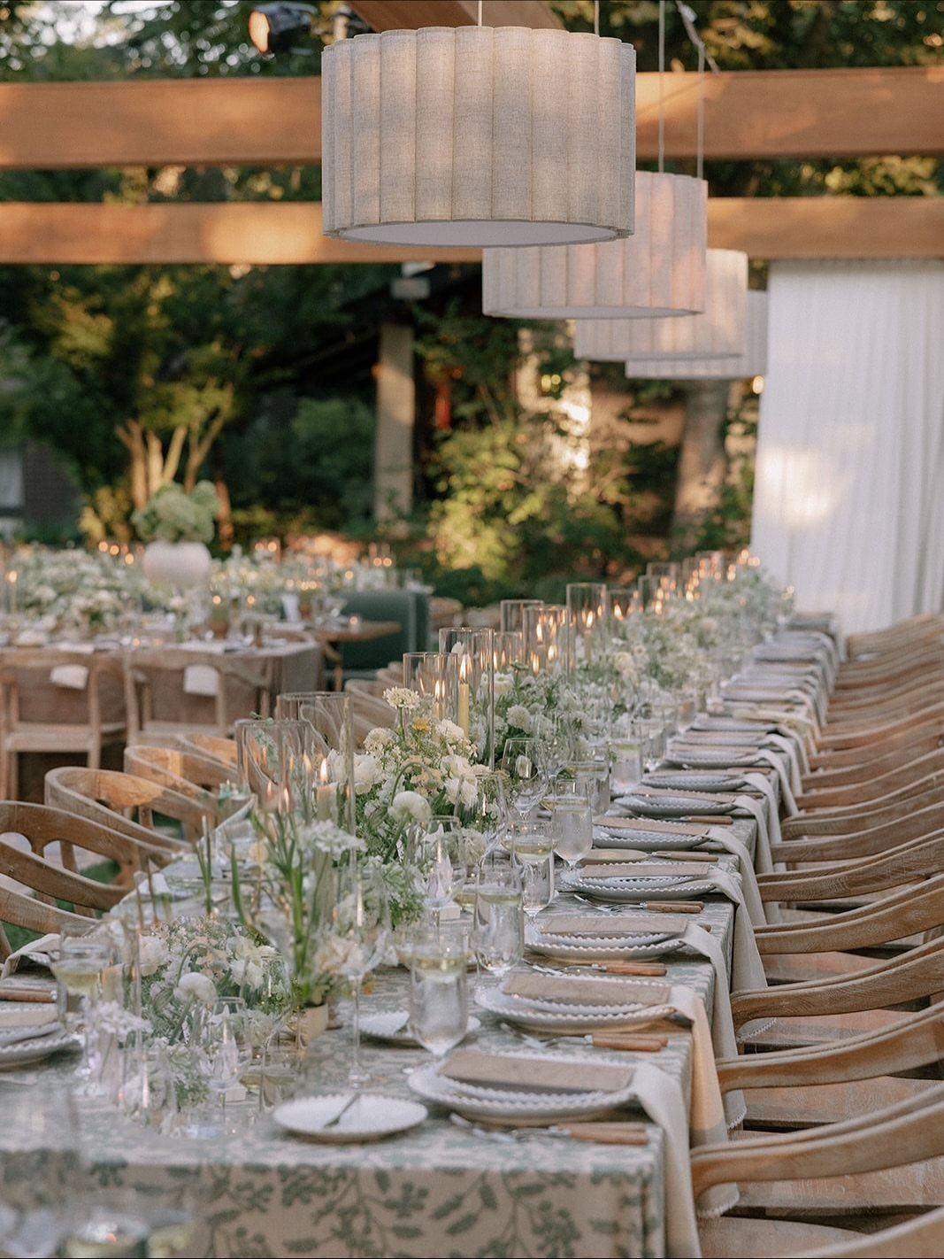 Long outdoor wedding table with white flowers and pendant lights under a pergola
