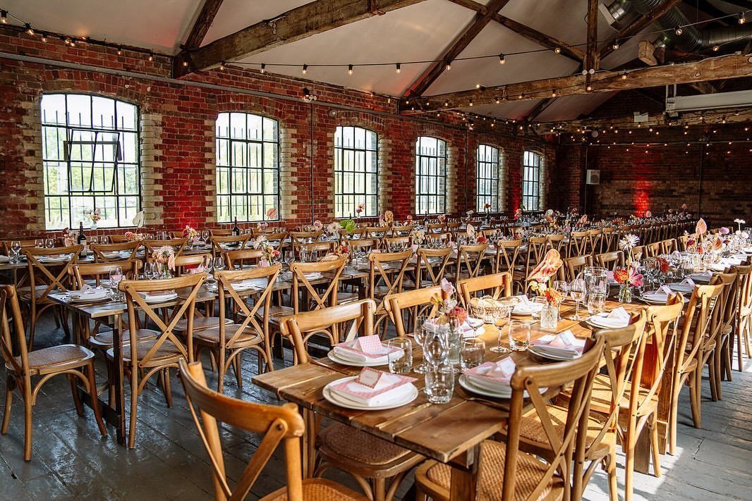 Industrial event space with red brick walls, wooden tables, and cross-back chairs.