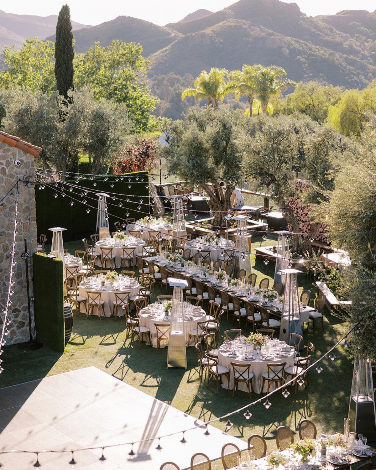 Outdoor wedding reception with white tables and chairs set among olive trees and mountains.