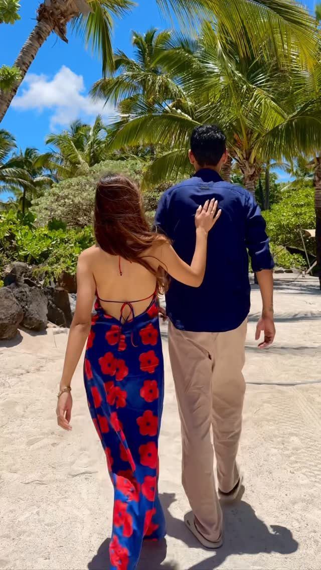 Couple walking on a sandy beach surrounded by palm trees under a blue sky.