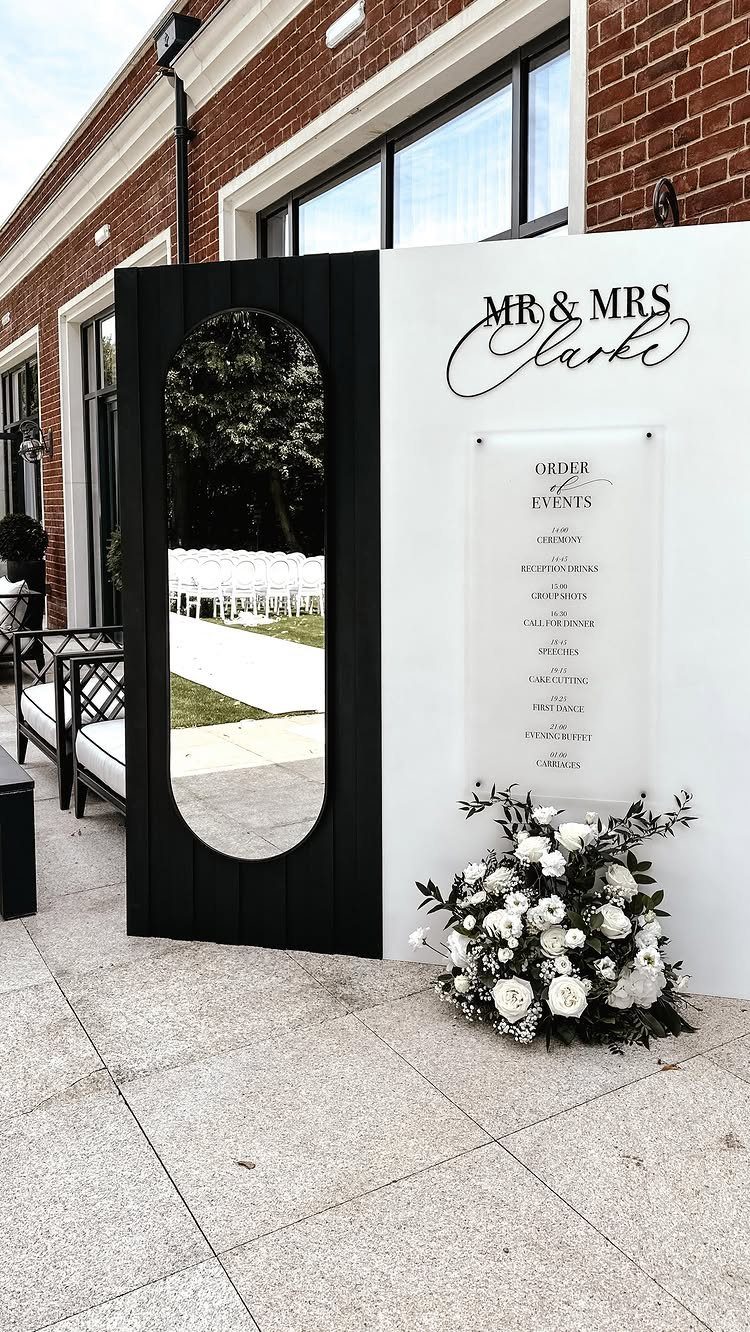 Wedding welcome display with custom signs, an oval mirror, and white flowers.