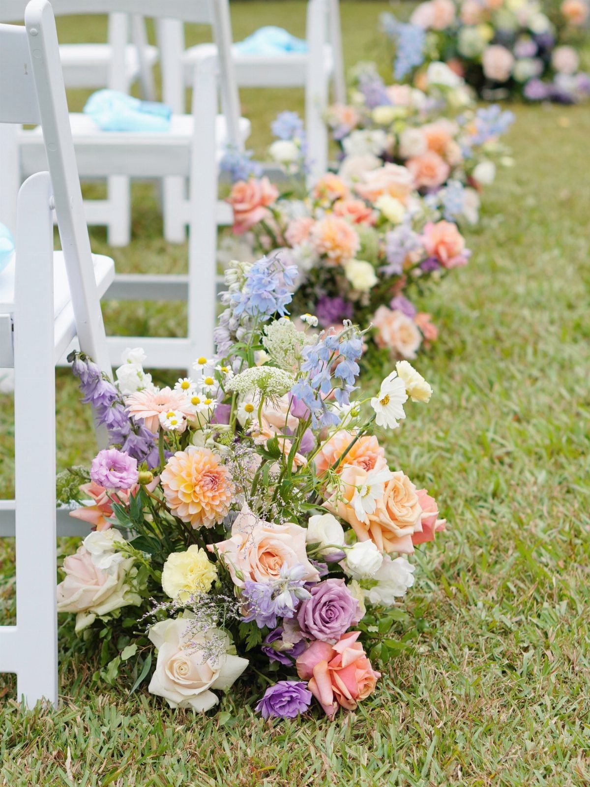 Pastel floral arrangements lining a grassy aisle next to white wedding chairs.