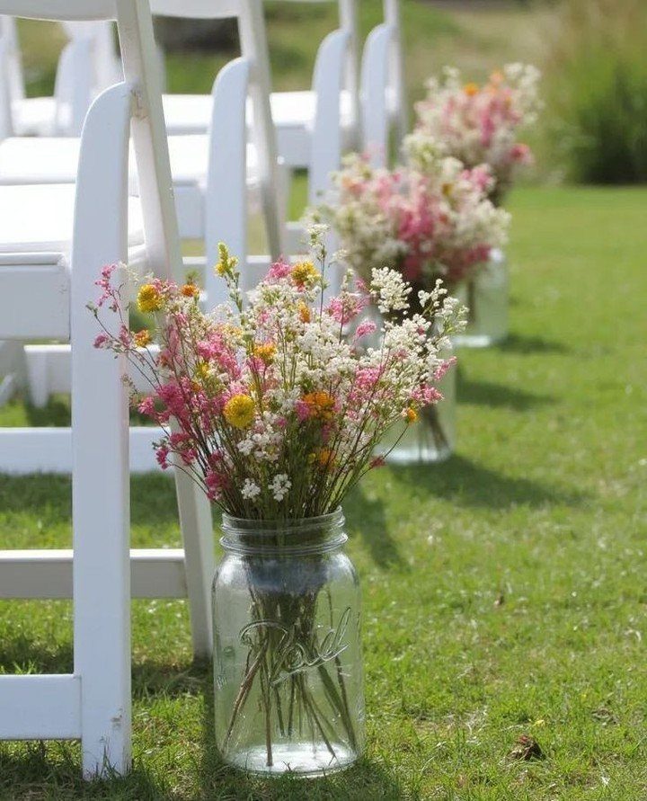 Wildflowers in mason jars lining a row of white chairs on green grass.