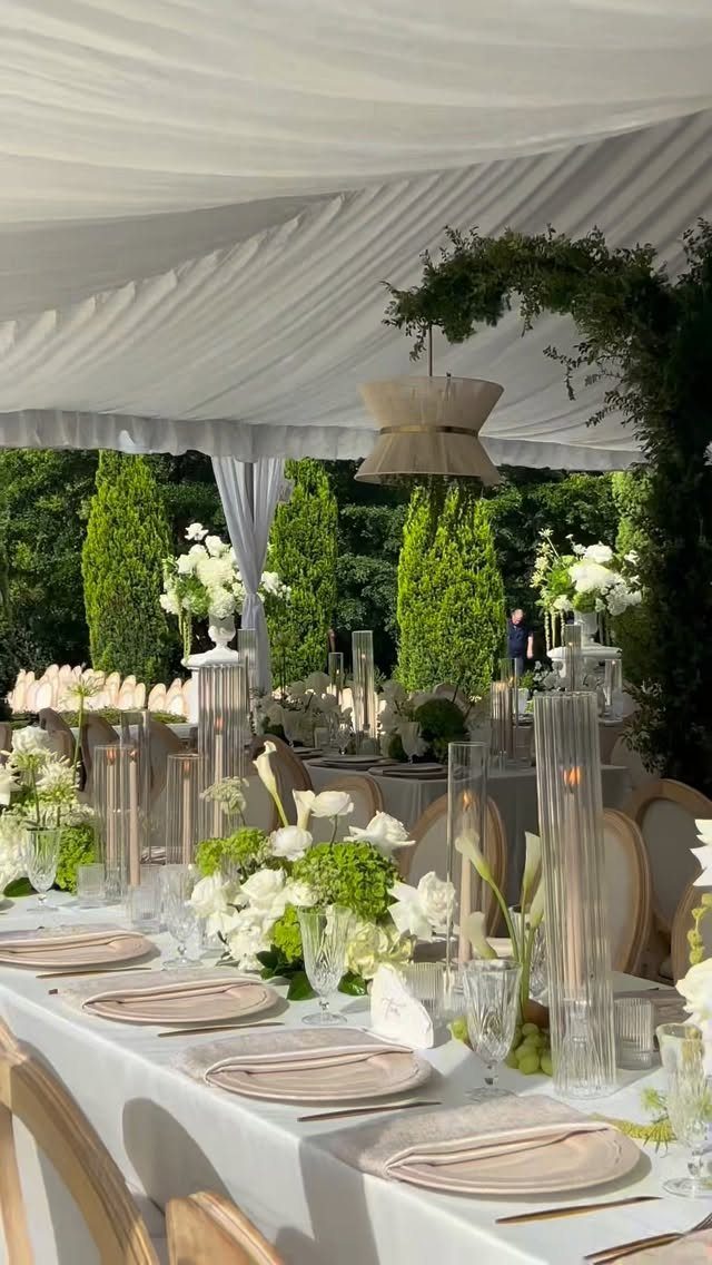 White floral arrangements and candles on a long table under a white wedding tent.