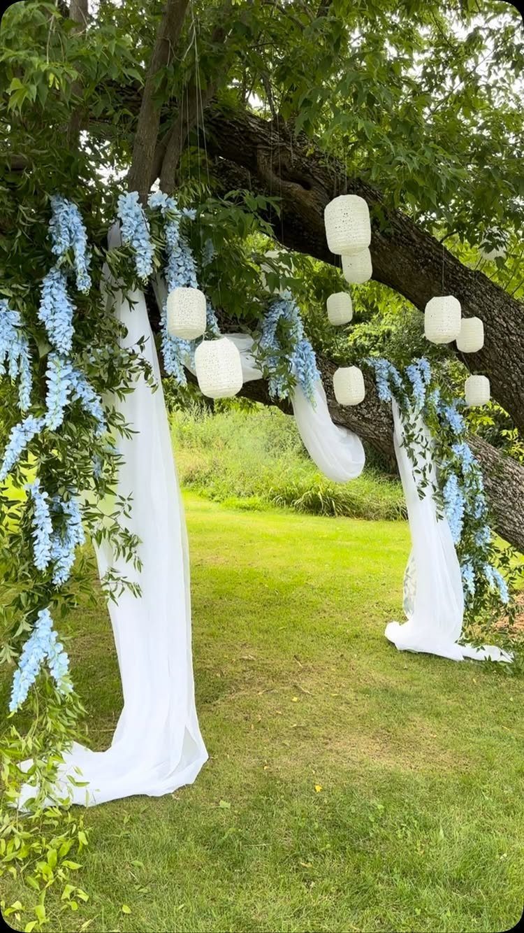 White paper lanterns and blue floral garlands hanging from a tree with white fabric.