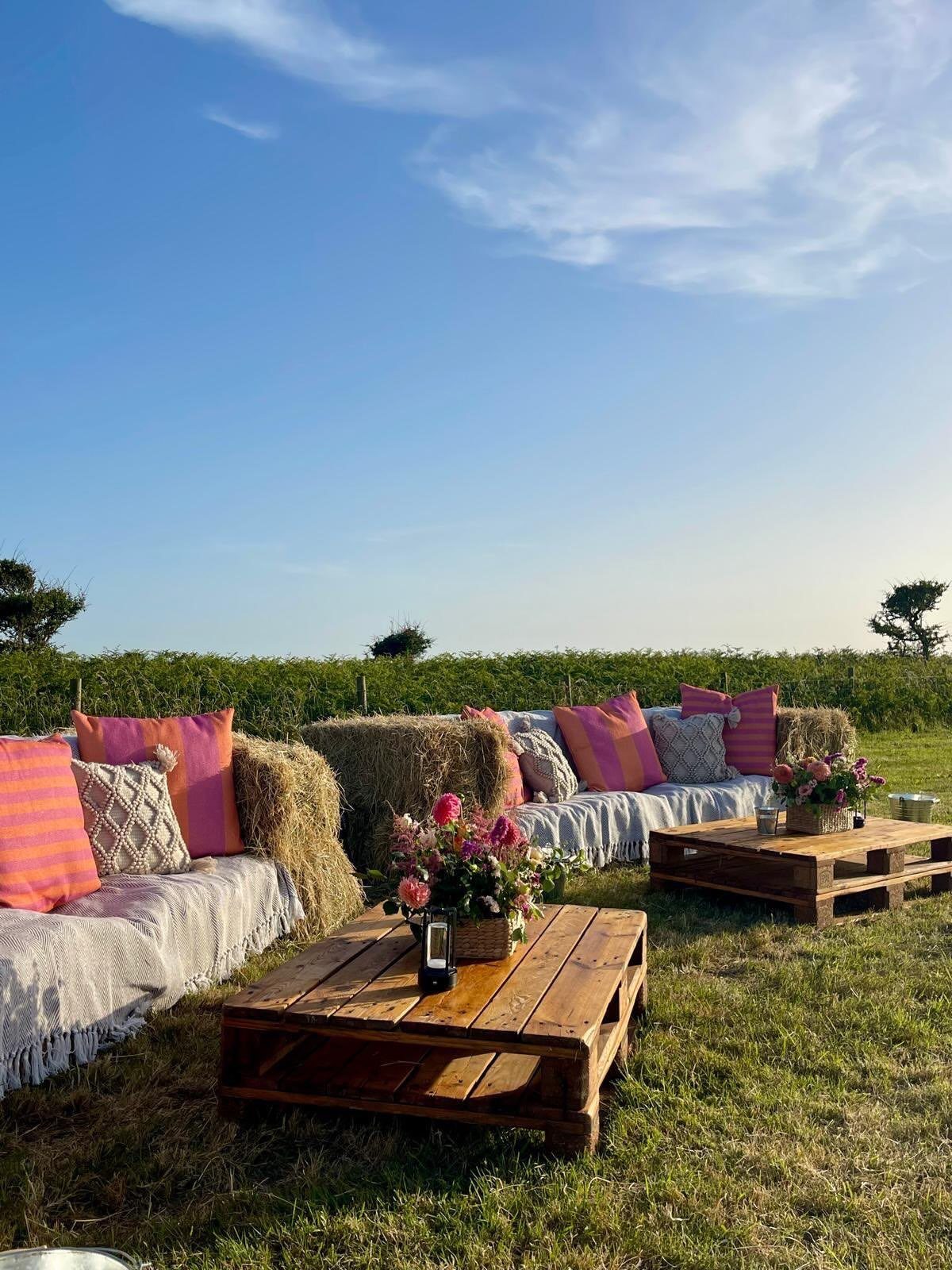 Hay bale seating with pink pillows and wooden pallet tables in a field.