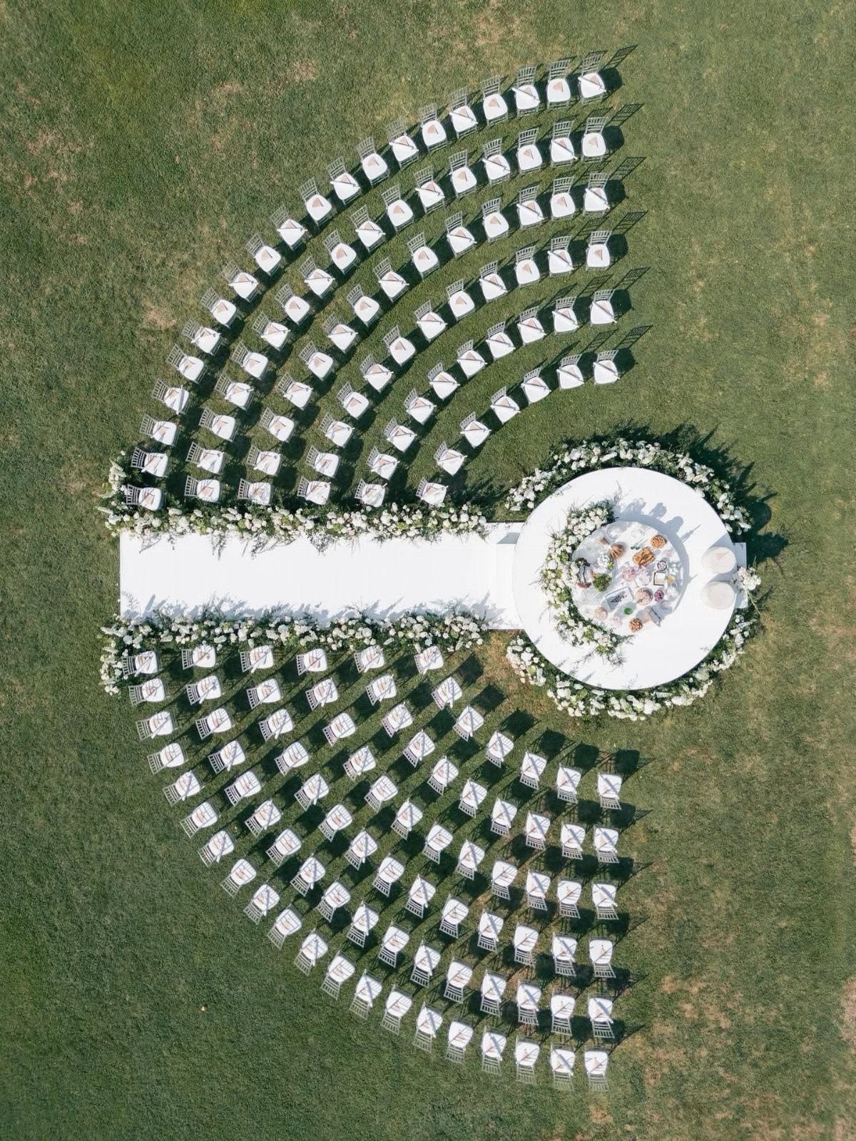 Curved rows of white chairs facing a circular floral altar on green grass.
