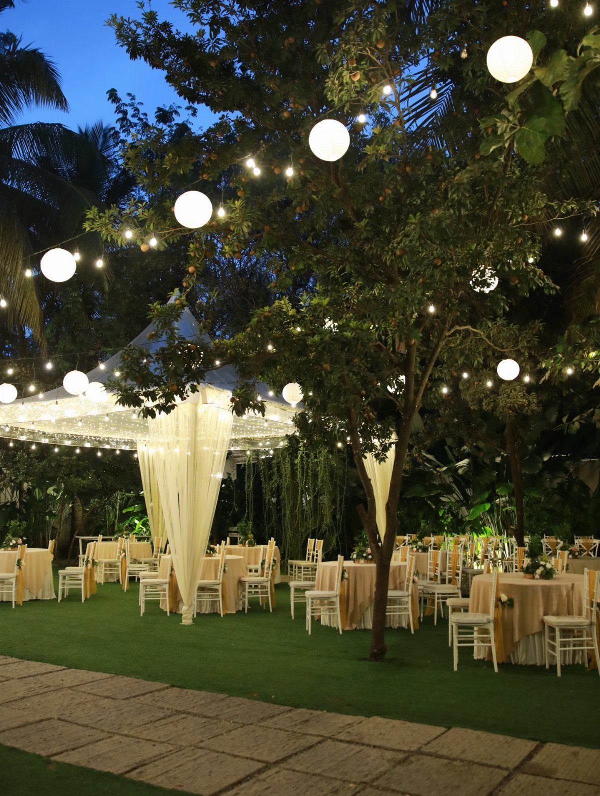 Outdoor event reception with beige tables, white chairs, and glowing globe lights in trees.