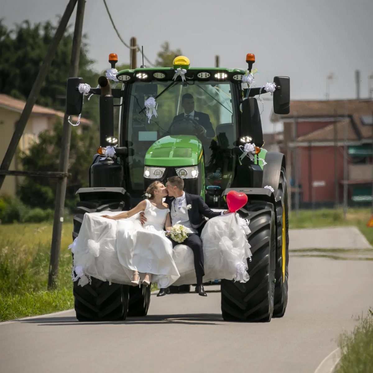 Bride and groom kissing on the front of a decorated green tractor on a road.