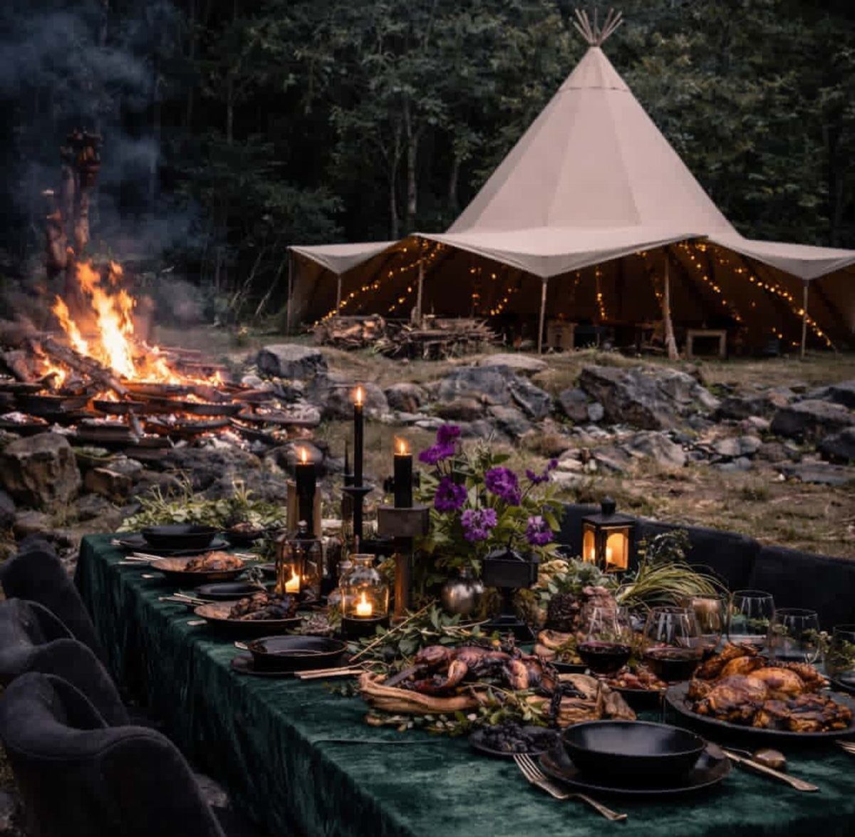 Outdoor dining table with green velvet cloth near a campfire and lit teepee.