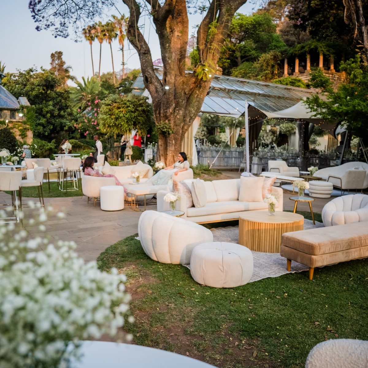 White curved sofas and round ottomans arranged as lounge seating outdoors under trees.