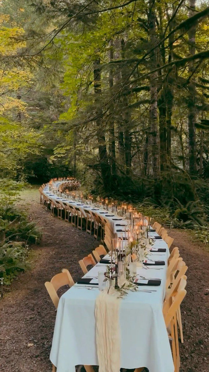 Long winding dining table with white linens and candles in a forest
