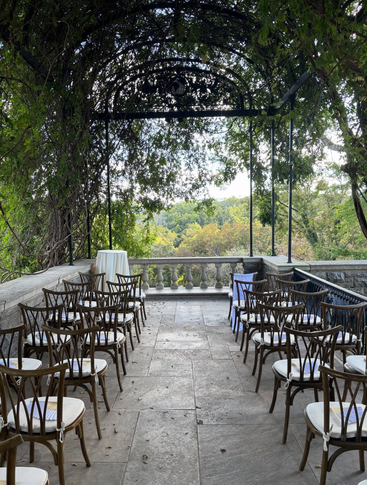 Wooden wedding chairs on a stone terrace under a vine-covered metal arbor.