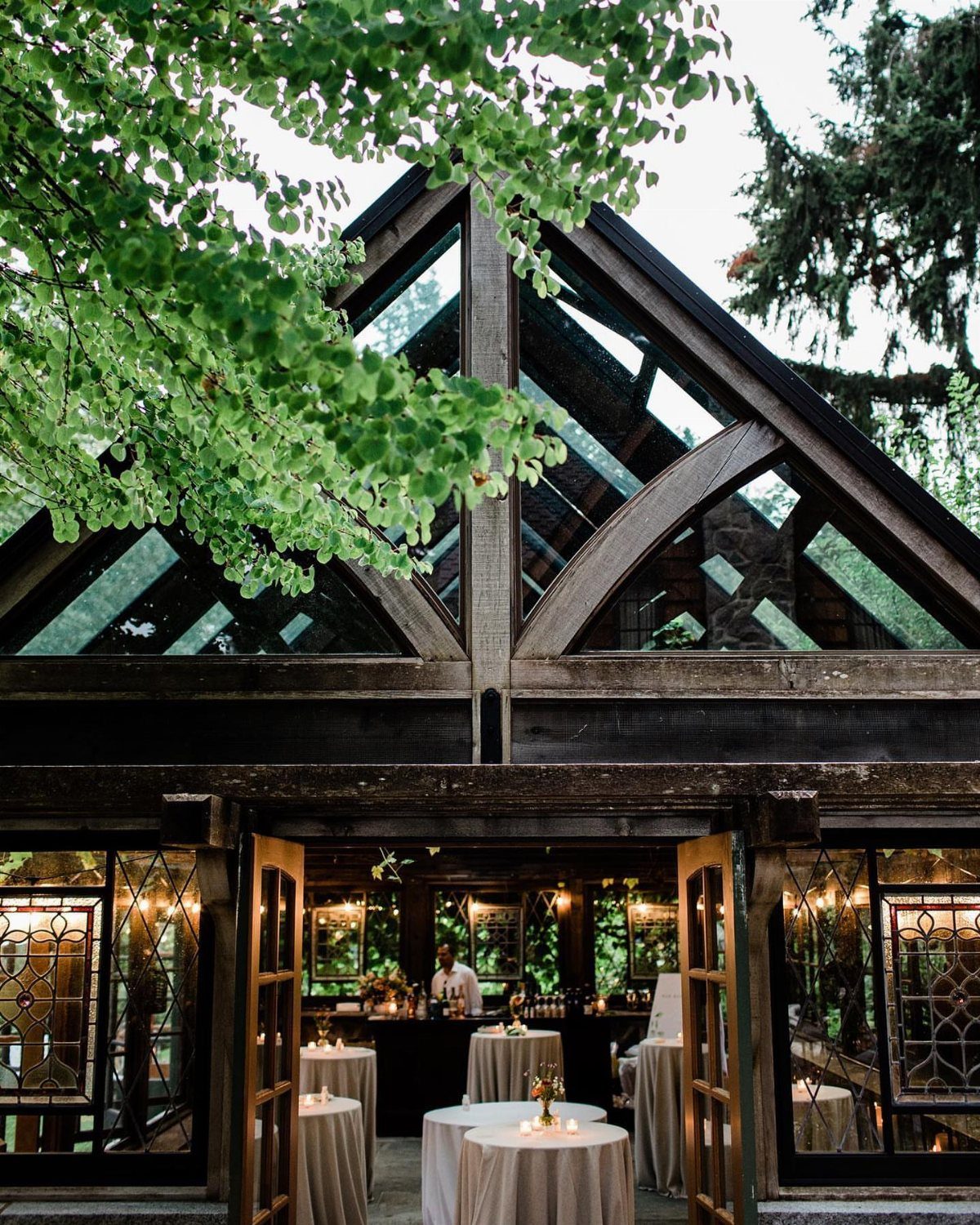 Glass greenhouse wedding venue with wooden beams and white cocktail tables inside.