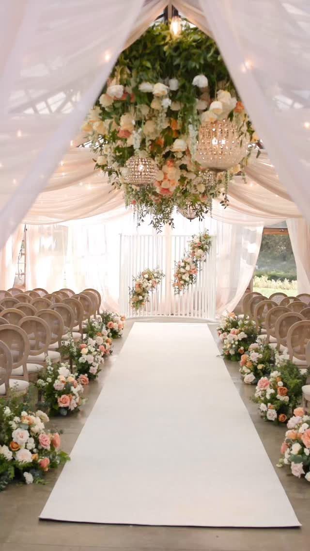 Wedding ceremony aisle with white runner, tan chairs, and hanging floral chandeliers.