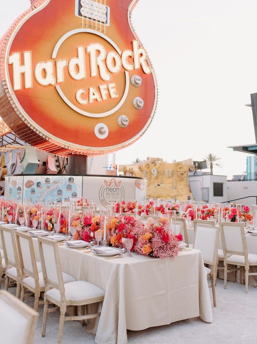 Outdoor wedding reception table with red flowers under a giant neon guitar sign.