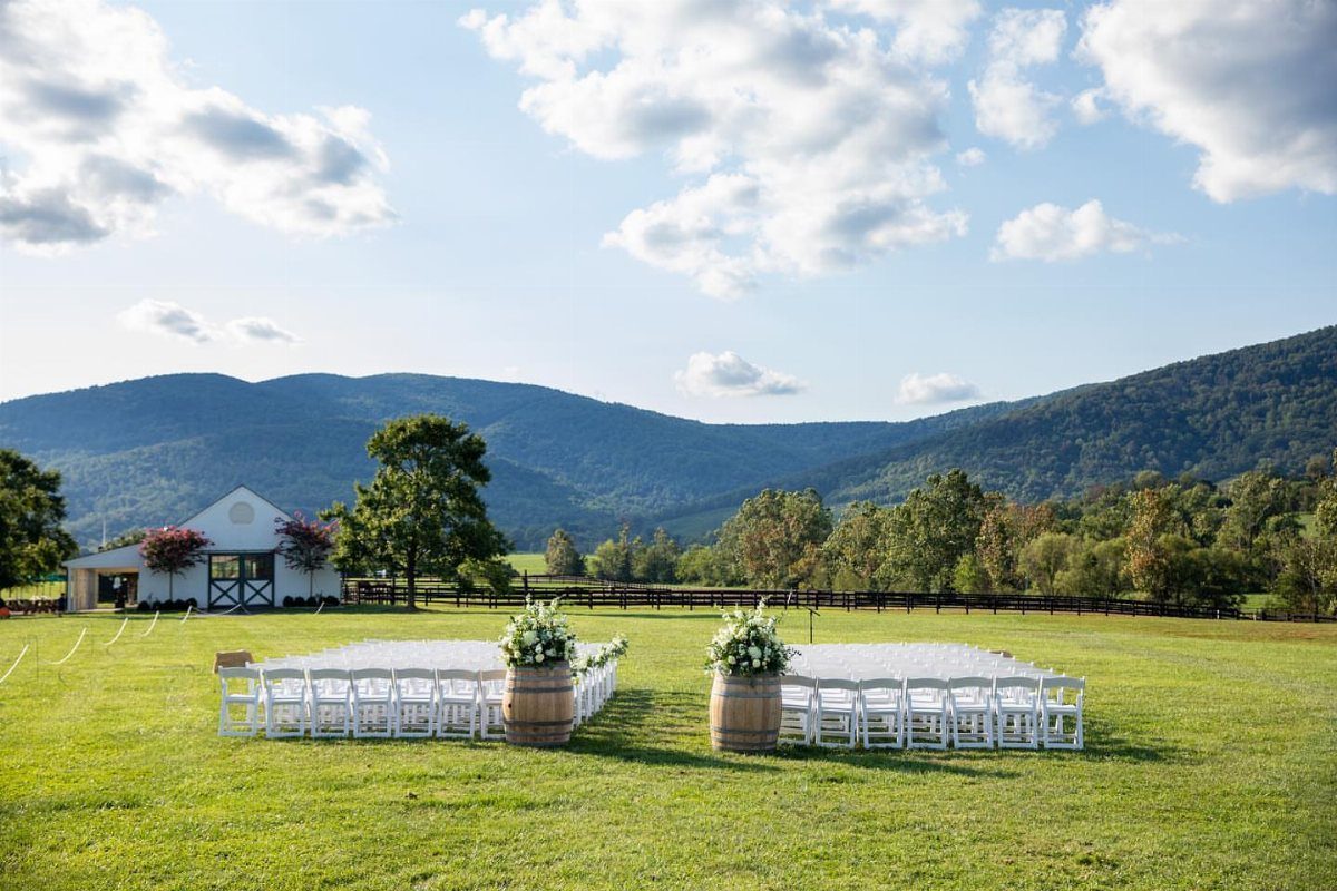 Outdoor wedding ceremony setup with white chairs and floral wine barrels before a barn.