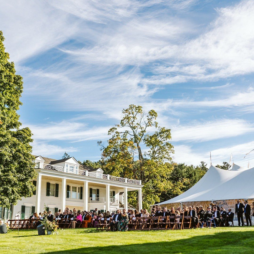 Wedding guests seated on a green lawn beside a white house and large tent.