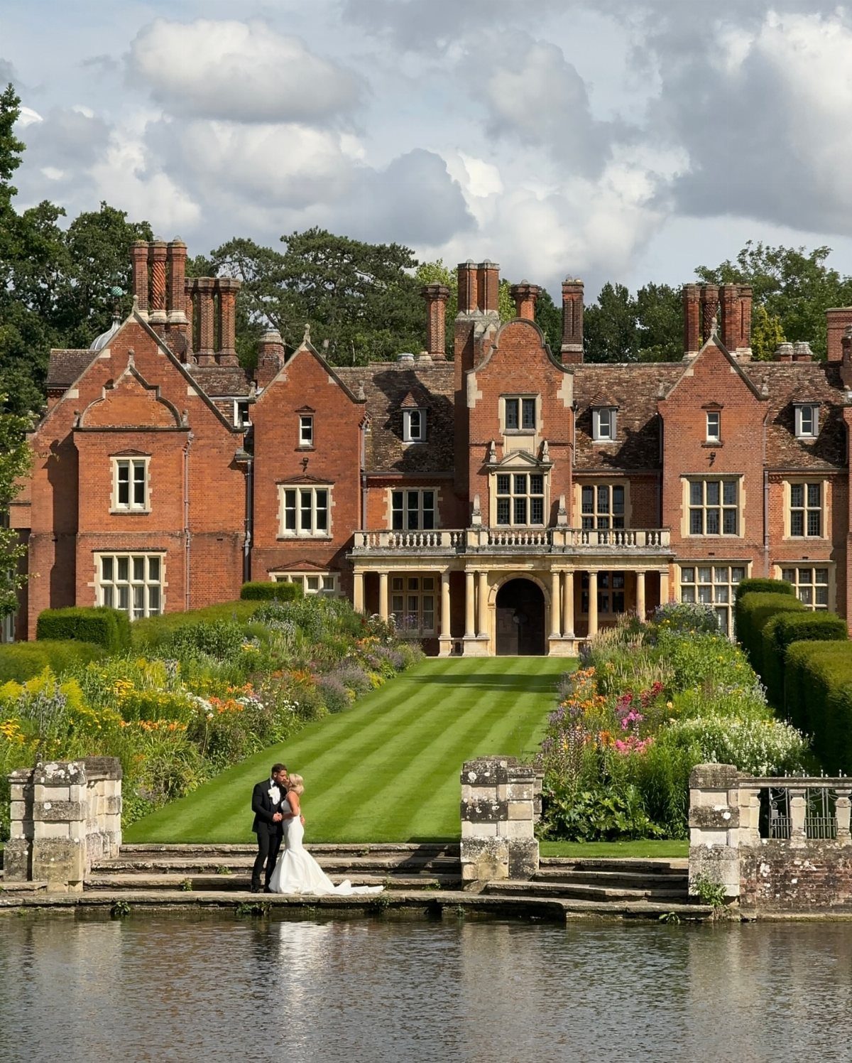 Bride and groom embracing by a pond before a large red-brick manor house.