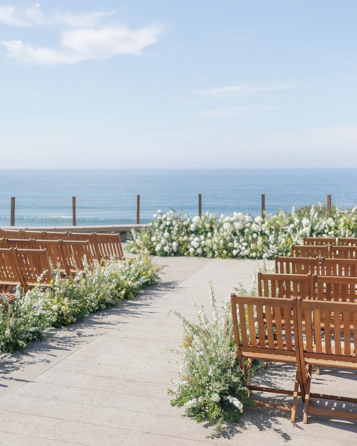 Wooden wedding chairs and white floral arrangements on a patio overlooking the ocean.