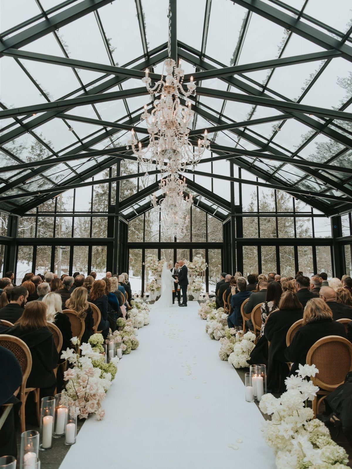 Wedding ceremony in a glass conservatory with a white aisle and crystal chandelier.