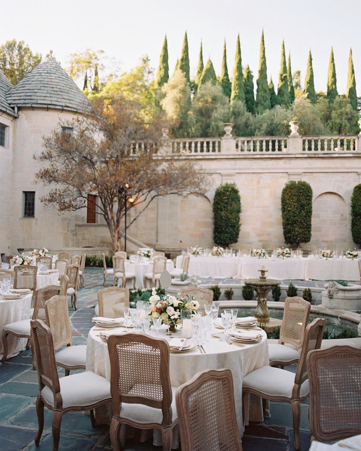 Outdoor wedding reception tables in a stone courtyard with a mansion and cypress trees.