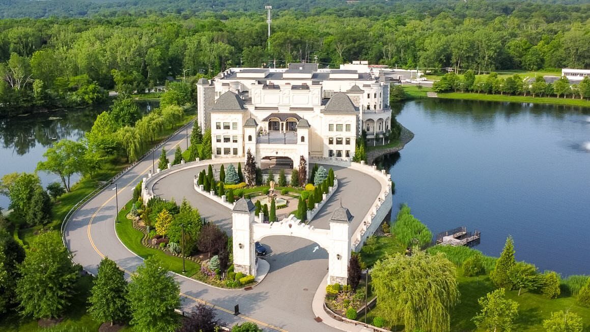 Large white luxury castle estate surrounded by a blue lake and green forest.